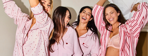 A Group of 4 women of varying ages wearing pink and red pyjamas with hearts and a fluffy robe standing on a bed like a PJ party