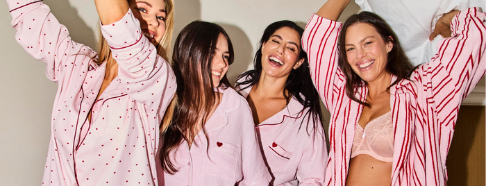 A Group of 4 women of varying ages wearing pink and red pyjamas with hearts and a fluffy robe standing on a bed like a PJ party