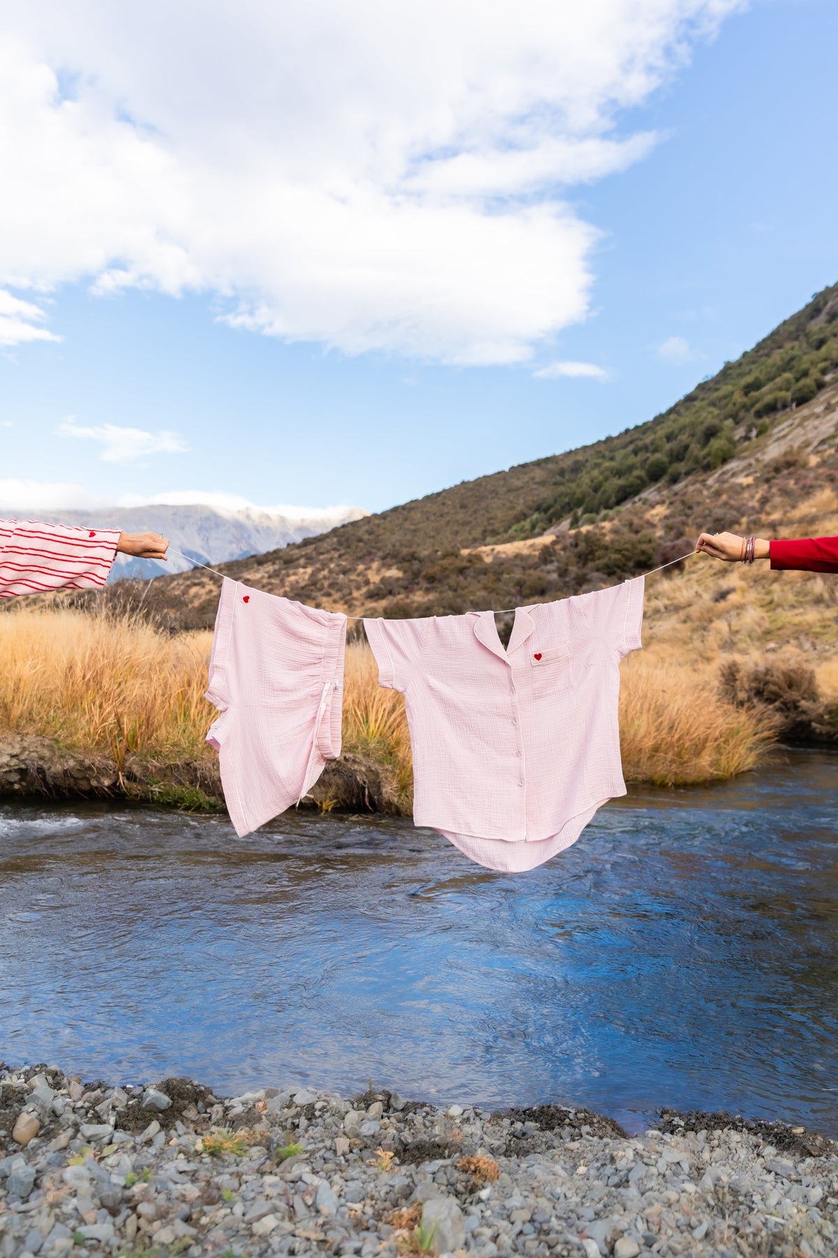 Two light pink shirts hanging on a string line outdoors by a river, with a mountain landscape, blue sky, and grassy bank in the background. A person's arms are visible on both sides holding the line.