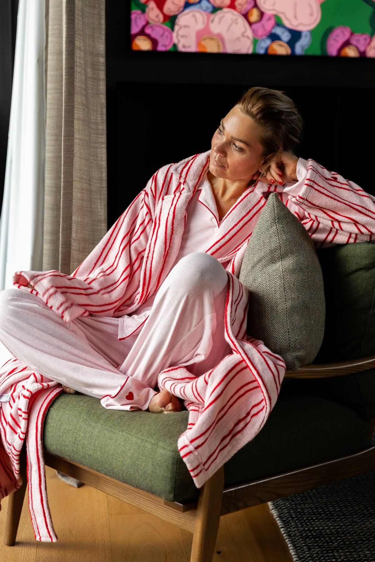 A woman wearing a pink pajama set with red and white striped robe and matching pants, sitting in a green armchair in a modern room.