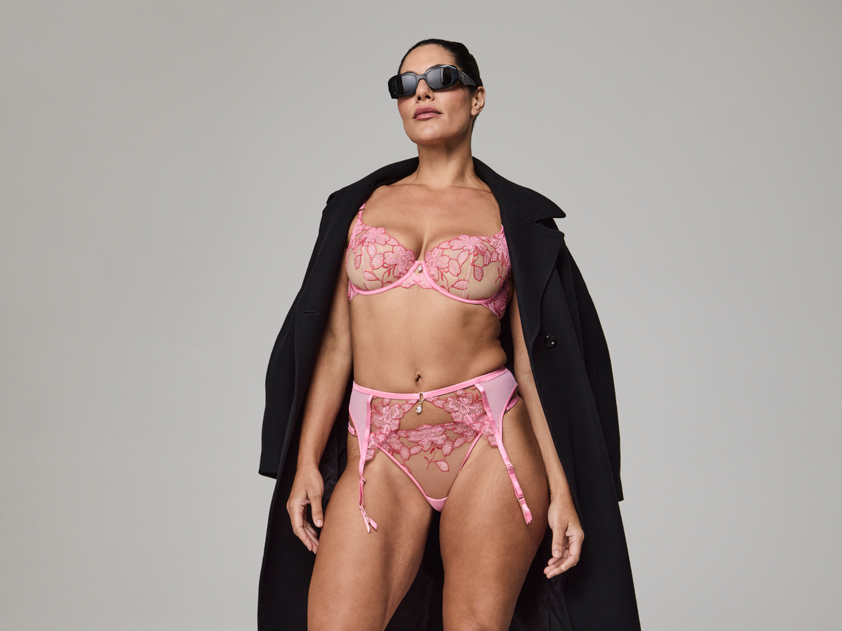 Woman wearing a pink lingerie set with sunglasses and a black coat on a gray background