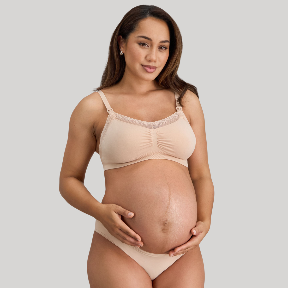 Pregnant woman wearing a beige maternity bra and underwear set on a gray background